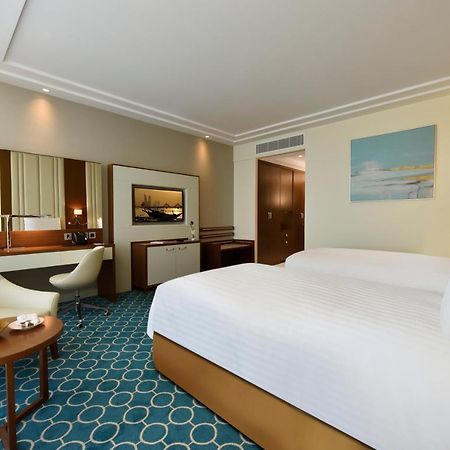 Grand Swiss-Belhotel Waterfront Seef Sanabis Esterno foto The photo shows a well-decorated hotel room featuring two beds with white linens. There's a comfortable seating area with a small round table that holds a bowl of fruit. A stylish chair is positioned next to the table. The room has a modern design wi