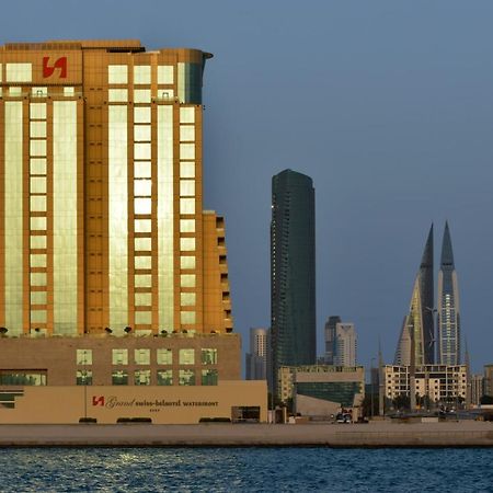 Grand Swiss-Belhotel Waterfront Seef Sanabis Esterno foto The image shows a waterfront cityscape featuring several buildings. In the foreground, there is a tall building with a golden exterior, reflecting sunlight. This building appears to be modern and is likely used for commercial purposes. Behind it, the