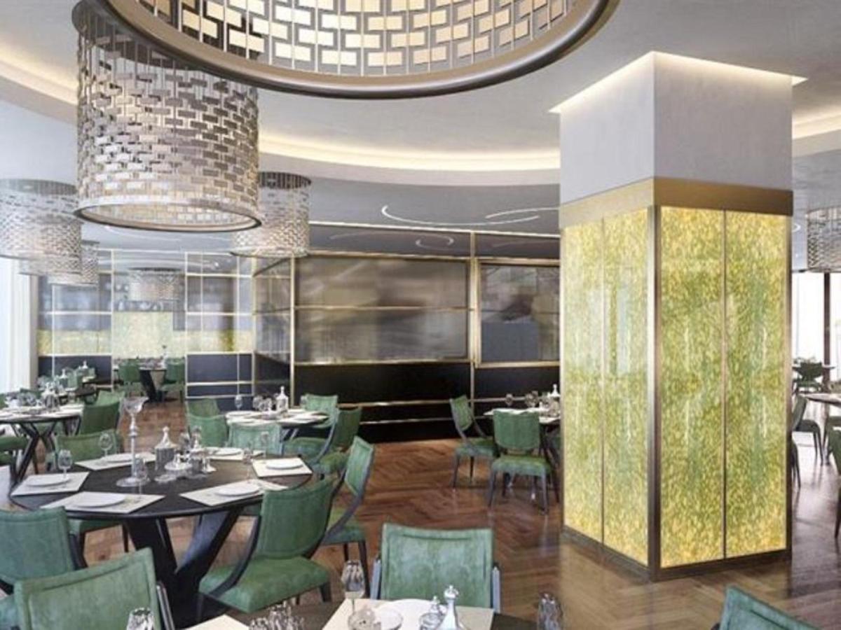 Grand Swiss-Belhotel Waterfront Seef Sanabis Esterno foto The photo shows the interior of a modern restaurant. It features a sophisticated design with stylish, green upholstered chairs arranged around dark tables. The dining area is illuminated by large, elegant light fixtures hanging from the ceiling. Ther