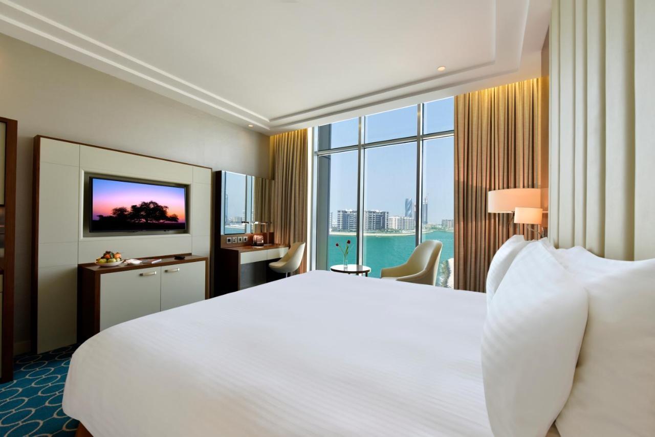 Grand Swiss-Belhotel Waterfront Seef Sanabis Esterno foto The image shows a modern hotel room featuring a large, neatly made bed with white bedding. There is a wall-mounted television opposite the bed, and a small desk with a chair is situated near large windows that appear to offer a view of the outside. T