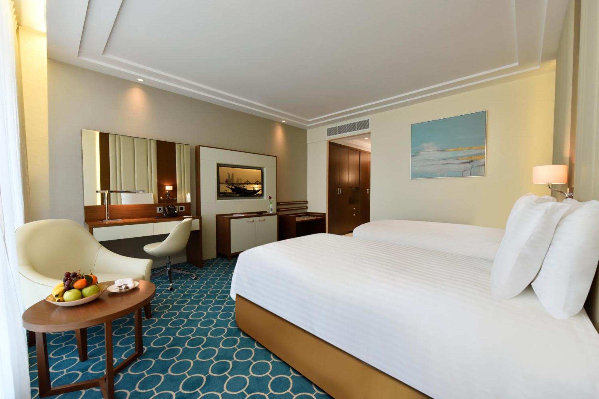 Grand Swiss-Belhotel Waterfront Seef Sanabis Esterno foto The photo shows a well-decorated hotel room featuring two beds with white linens. There's a comfortable seating area with a small round table that holds a bowl of fruit. A stylish chair is positioned next to the table. The room has a modern design wi