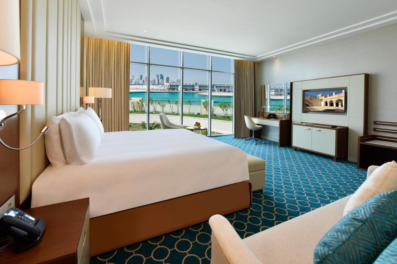 Grand Swiss-Belhotel Waterfront Seef Sanabis Esterno foto The photo shows a luxurious hotel room. It features a large bed with white linens, and there is a comfortable armchair and a small sofa in the corner. The room has large windows that offer a view of the outdoors, likely facing a body of water. The ca
