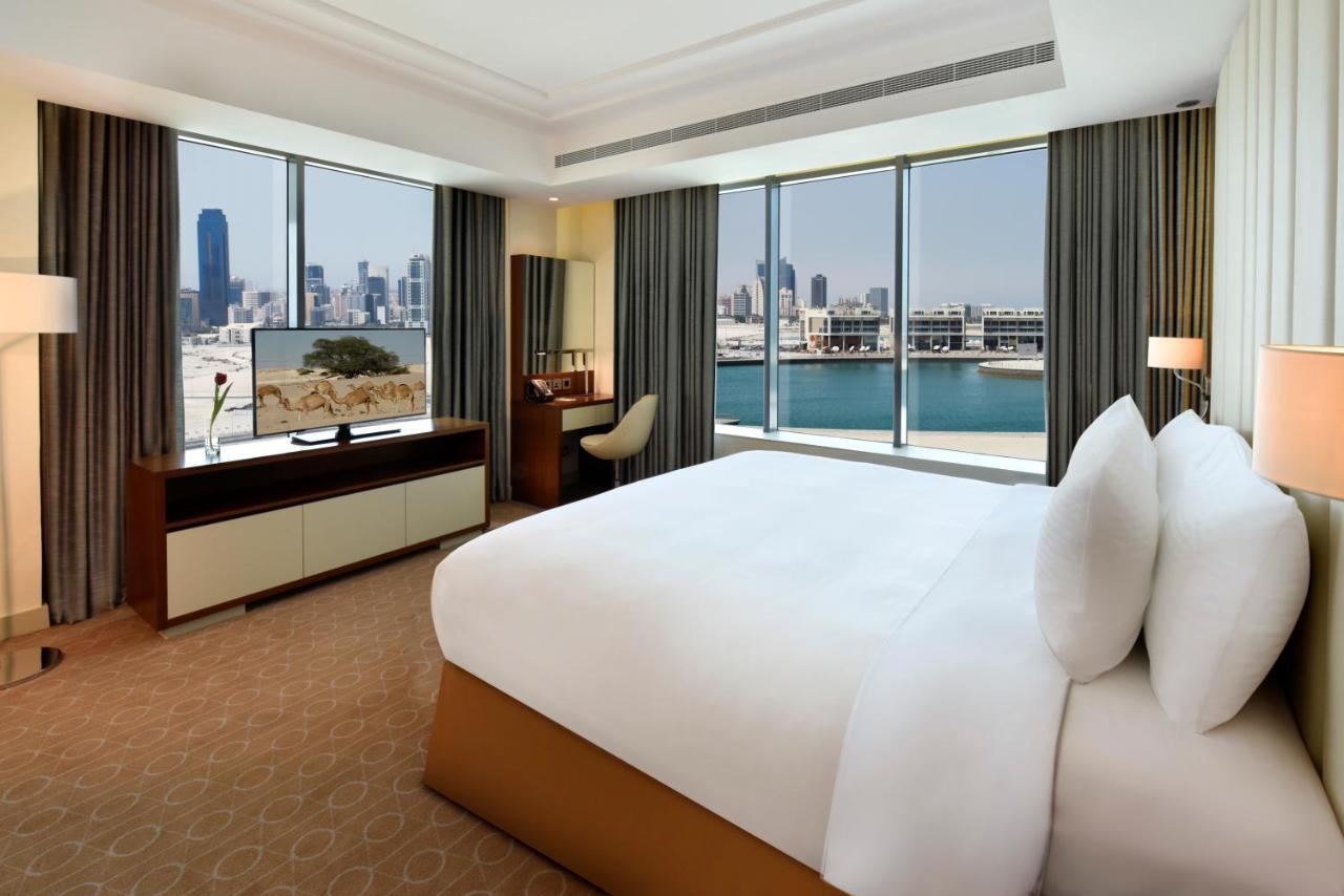 Grand Swiss-Belhotel Waterfront Seef Sanabis Esterno foto The photo shows a luxurious hotel room featuring a large bed with white pillows and a neatly made bedspread. The room has floor-to-ceiling windows that offer a view of a city skyline, suggesting a vibrant urban setting. There is a modern television m