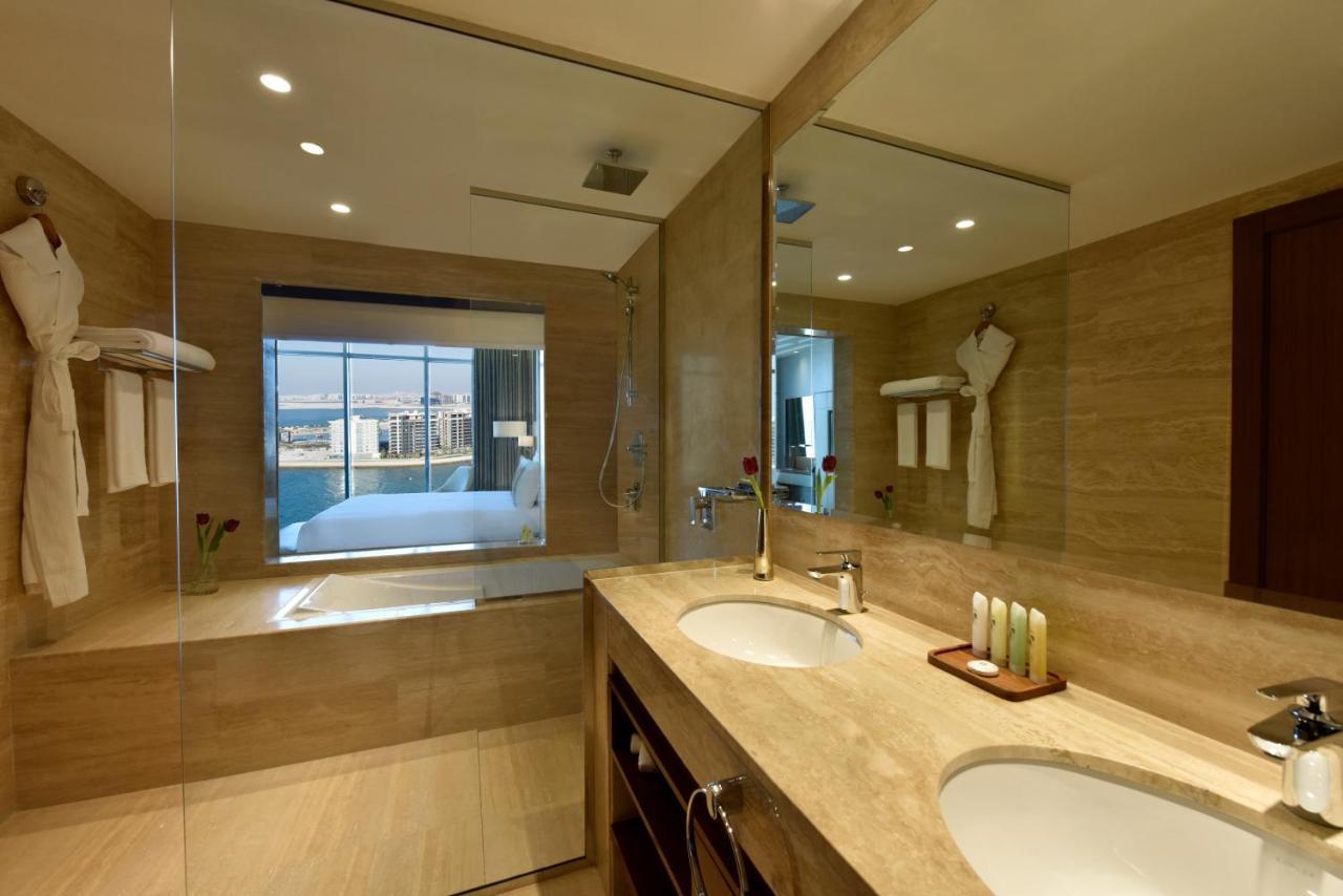 Grand Swiss-Belhotel Waterfront Seef Sanabis Esterno foto The photo depicts a modern bathroom with a sleek design. It features a large mirror above a double sink with a wooden cabinet below. The walls are adorned with light-colored tiles, and there is a glass shower enclosure. On one side of the room, there
