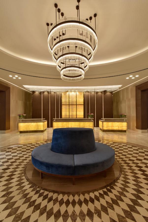 Grand Swiss-Belhotel Waterfront Seef Sanabis Esterno foto The photo shows an elegant hotel lobby. It features a circular seating area in the center with a plush, dark blue upholstered bench. Surrounding this seating area is a patterned floor made of light and dark tiles. Behind the seating area, there are t