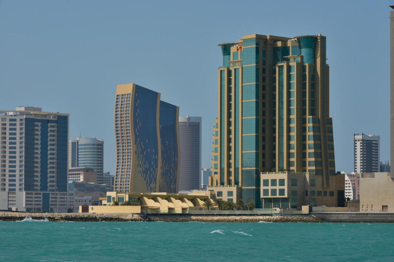 Grand Swiss-Belhotel Waterfront Seef Sanabis Esterno foto The photo showcases a coastal city skyline featuring several modern buildings. Prominent in the foreground is a tall structure with a distinctive design, characterized by a mix of green, gold, and glass. Next to it is another building with a more cur