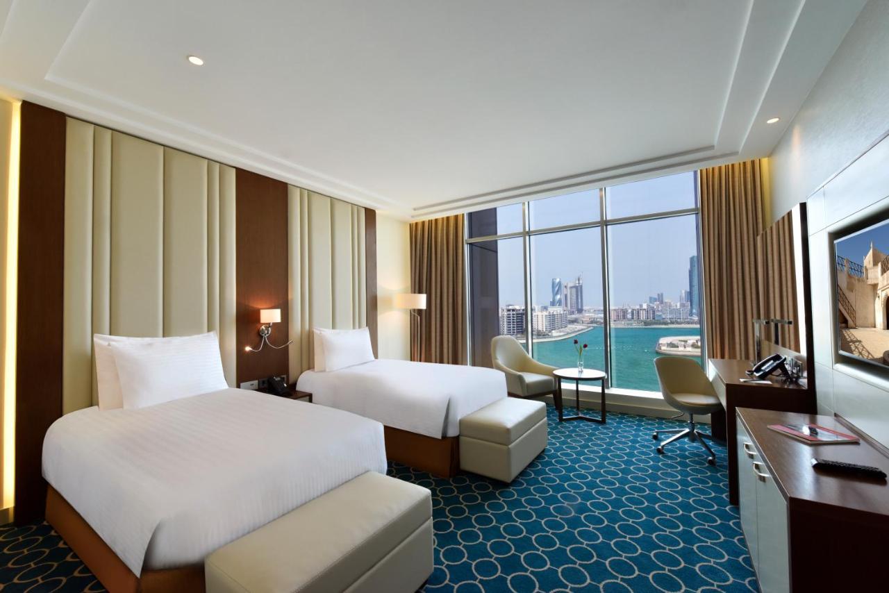 Grand Swiss-Belhotel Waterfront Seef Sanabis Esterno foto The photo shows a hotel room featuring two twin beds with white bedding. The interior design includes soft, neutral tones on the walls, accented by a textured panel. There are large windows that offer a view of a body of water and city skyline in the