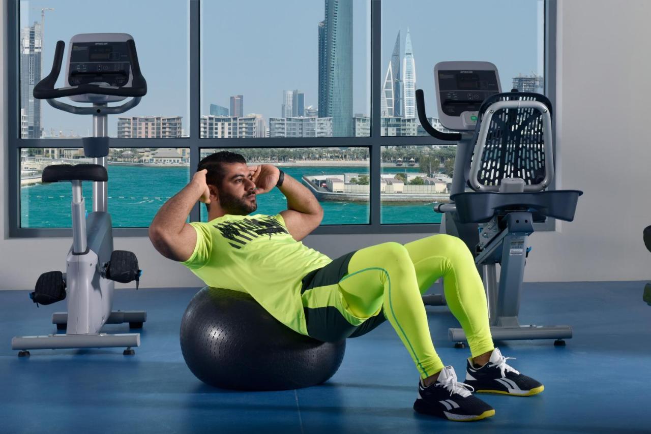 Grand Swiss-Belhotel Waterfront Seef Sanabis Esterno foto The photo shows a man exercising in a gym setting. He is performing a sit-up while balancing on an exercise ball. He is wearing a bright neon yellow athletic outfit and black shoes. In the background, large windows provide a view of a city skyline an