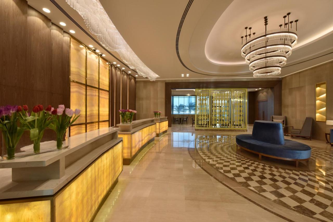 Grand Swiss-Belhotel Waterfront Seef Sanabis Esterno foto The photo shows an elegant hotel or lobby area. There is a reception desk with a warm yellow glow, flanked by potted flowers, possibly tulips. The interior features a modern design with a sophisticated chandelier hanging from the ceiling. To the side