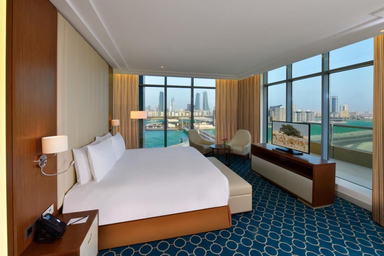 Grand Swiss-Belhotel Waterfront Seef Sanabis Esterno foto The photo shows a modern hotel room featuring a large bed with white linens positioned against a wall. There are large windows offering a view of the city and water beyond, allowing natural light to fill the space. The room includes a small seating a