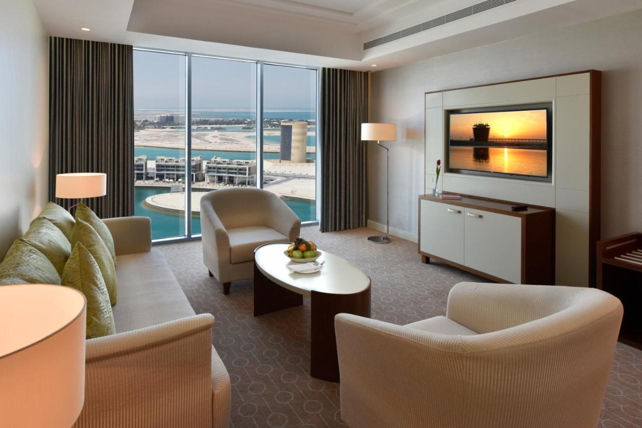 Grand Swiss-Belhotel Waterfront Seef Sanabis Esterno foto The photo depicts a luxurious hotel living room. It features a comfortable seating area with a light-colored sofa and armchairs, a stylish coffee table, and decorative elements like a fruit bowl. The large windows offer a stunning view of a body of w