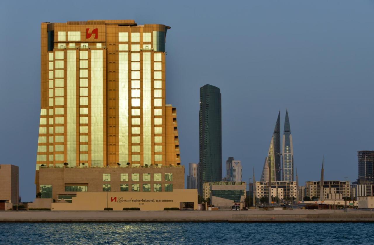Grand Swiss-Belhotel Waterfront Seef Sanabis Esterno foto The image shows a waterfront cityscape featuring several buildings. In the foreground, there is a tall building with a golden exterior, reflecting sunlight. This building appears to be modern and is likely used for commercial purposes. Behind it, the