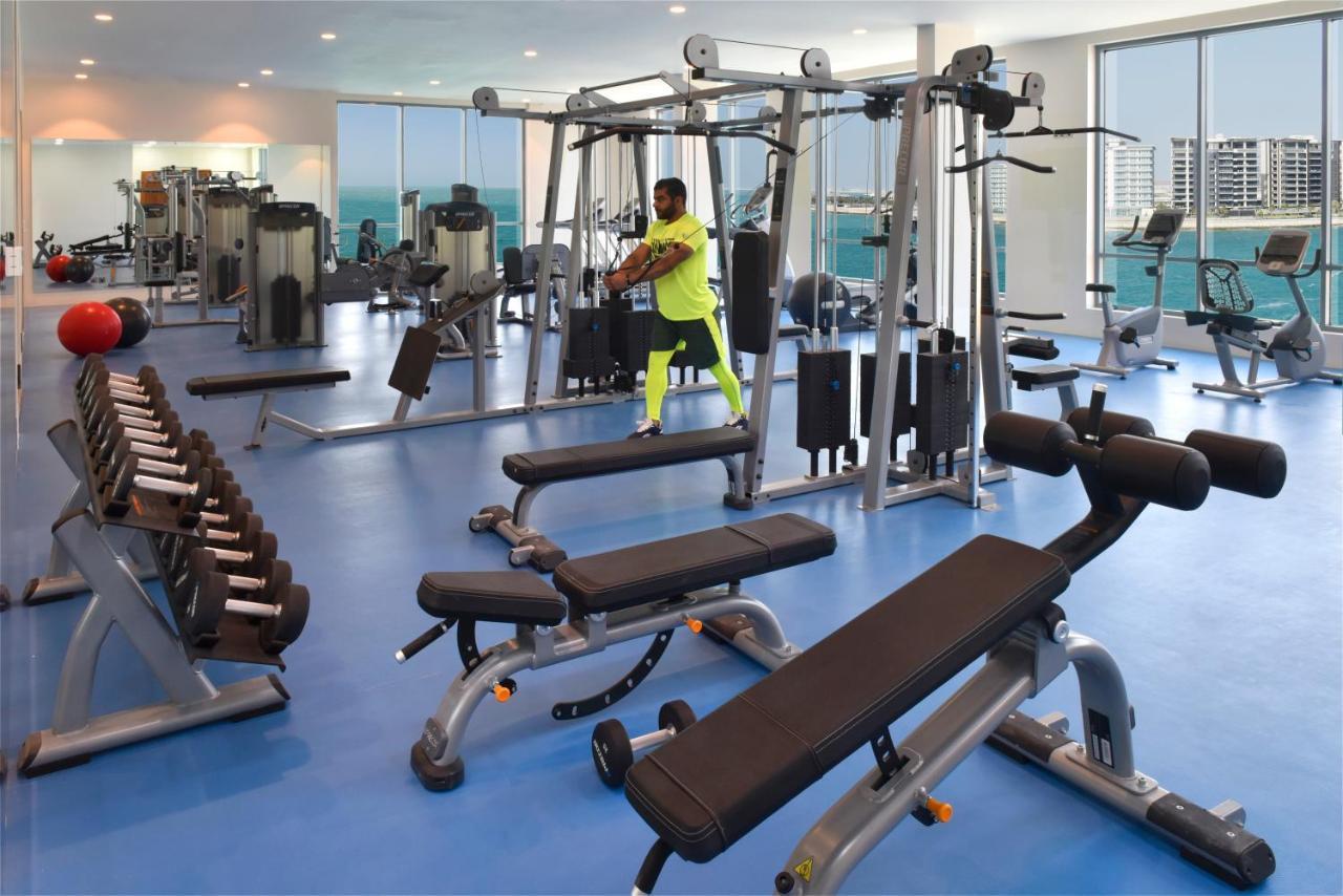 Grand Swiss-Belhotel Waterfront Seef Sanabis Esterno foto The photo shows a modern gym interior with a variety of fitness equipment. In the foreground, there is a weight bench and a set of dumbbells arranged neatly. To one side, there is a multi-station workout machine where a person is performing an exerci