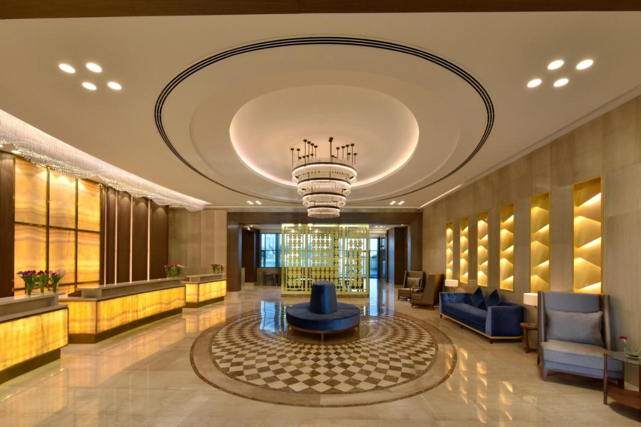 Grand Swiss-Belhotel Waterfront Seef Sanabis Esterno foto The image depicts a modern hotel lobby. It features a stylish design with warm lighting and elegant decor. There is a reception desk made of light-colored wood and illuminated panels. The ceiling has a large, circular light fixture, adding a luxuriou
