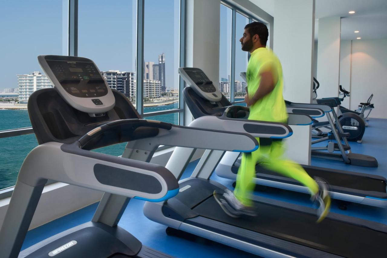 Grand Swiss-Belhotel Waterfront Seef Sanabis Esterno foto The photo features a modern gym setting with several treadmills positioned next to large windows that showcase a cityscape and waterfront view. A man is running on one of the treadmills, wearing bright neon athletic attire. The overall atmosphere app