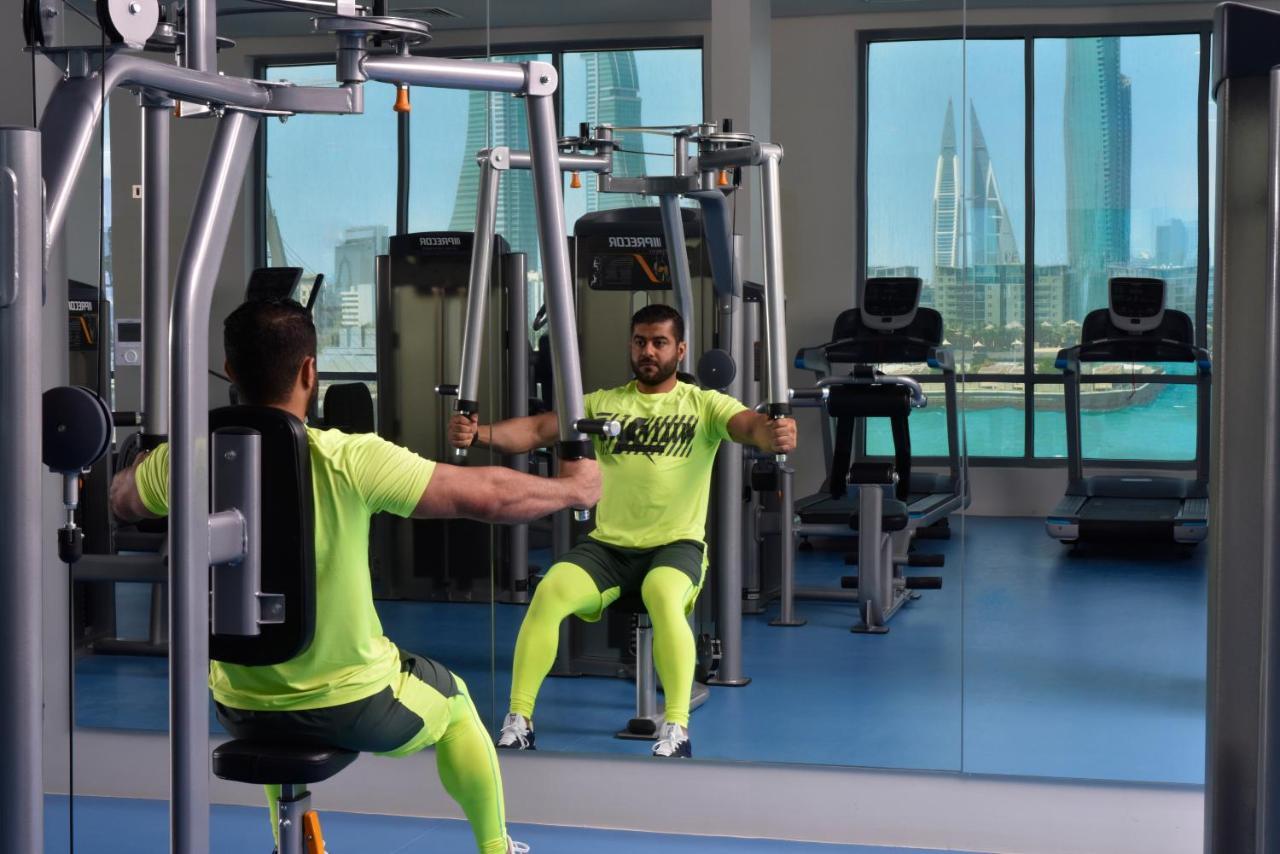 Grand Swiss-Belhotel Waterfront Seef Sanabis Esterno foto The photo shows a gym setting where a man is using a chest press machine. He is wearing bright yellow workout attire and is seated in front of a mirror that reflects his position. The gym has large windows that display a city skyline in the backgroun