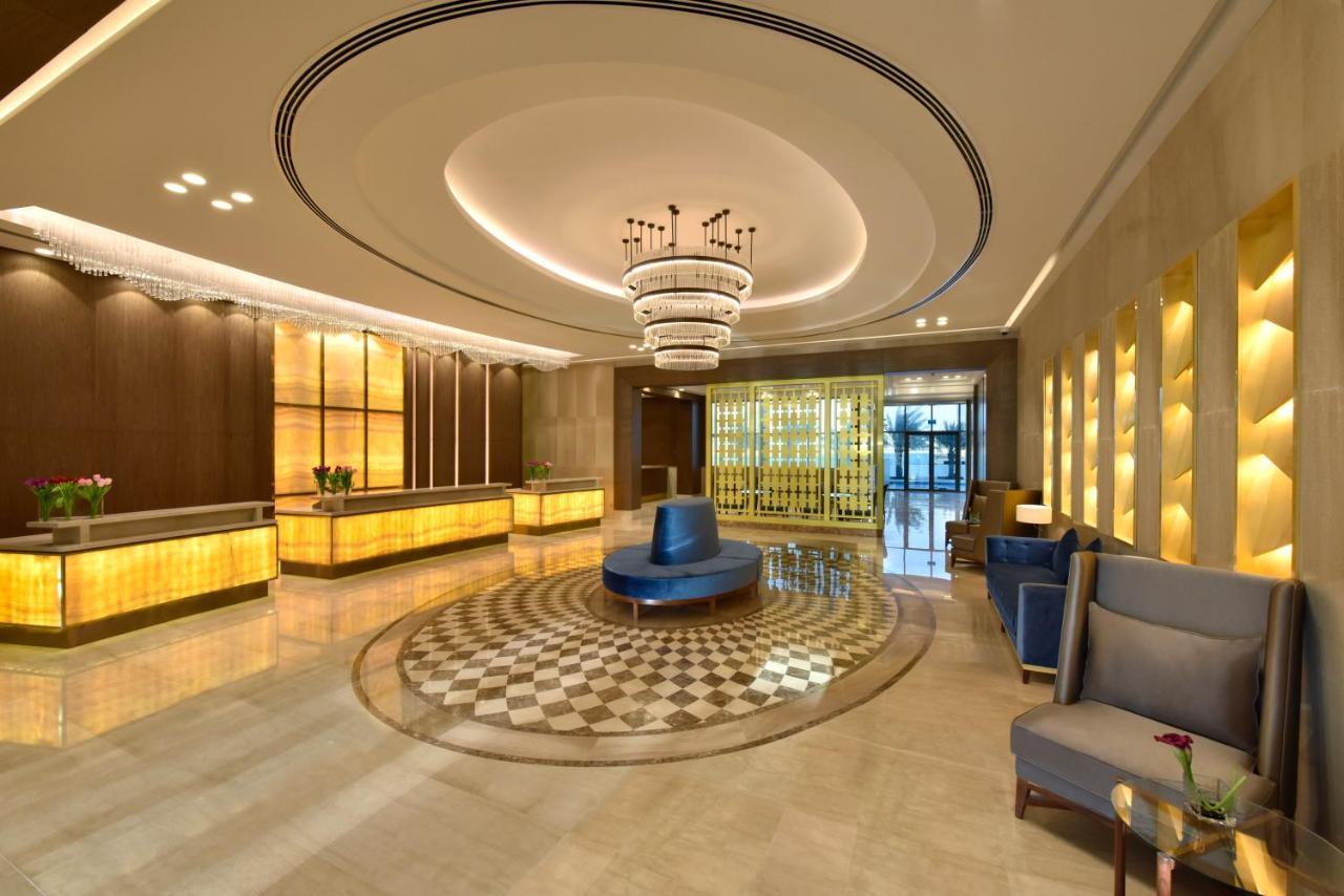 Grand Swiss-Belhotel Waterfront Seef Sanabis Esterno foto The image shows a modern hotel lobby. It features a spacious area with elegant design elements, including a large, decorative chandelier hanging from a curved ceiling. The reception desk is made of polished wood and illuminated by soft lighting. In t