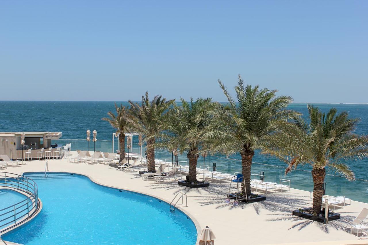 Grand Swiss-Belhotel Waterfront Seef Sanabis Esterno foto The photo depicts a scenic view of a tropical landscape with a swimming pool in the foreground. The pool has a curved shape and is surrounded by white lounge chairs. In the background, there are several tall palm trees providing shade. Beyond the poo