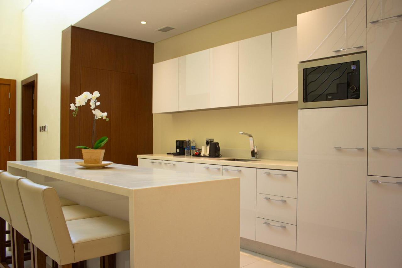 Grand Swiss-Belhotel Waterfront Seef Sanabis Esterno foto The photo shows a modern kitchen design featuring sleek white cabinets and countertops. On one side, there is a small island with bar stools, providing a space for seating. A potted plant, likely an orchid, is placed on the island. The kitchen is wel