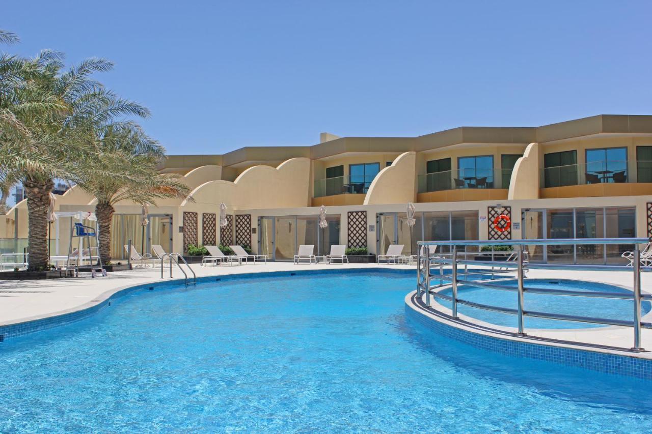 Grand Swiss-Belhotel Waterfront Seef Sanabis Esterno foto The photo shows a swimming pool with clear blue water, surrounded by a deck. There are lounge chairs arranged around the pool. In the background, there is a building with a modern architectural design featuring large windows and a light-colored facad