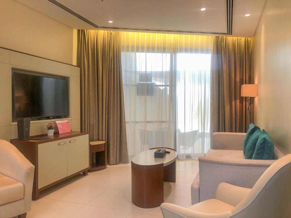 Grand Swiss-Belhotel Waterfront Seef Sanabis Esterno foto The photo shows a modern hotel room or a living space. It features a comfortable seating area with two white chairs and a small, round wooden coffee table. There is a flat-screen television mounted on the wall, with a console or cabinet below it. Lig