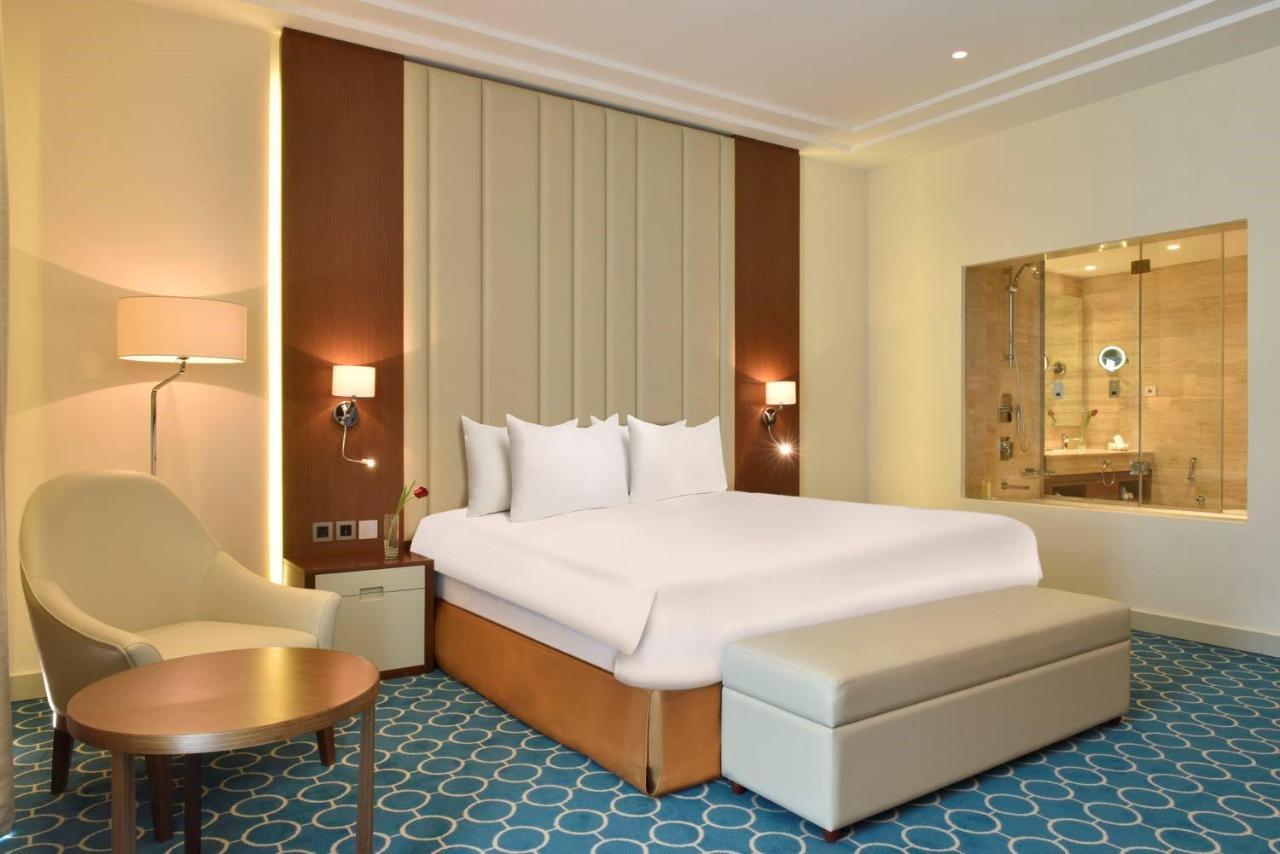 Grand Swiss-Belhotel Waterfront Seef Sanabis Esterno foto The photo shows a modern hotel room. It features a large bed with white bedding, placed against a wall with a textured design. To the side, there is a comfortable chair and a small round table. There is also a bench at the foot of the bed. The room h