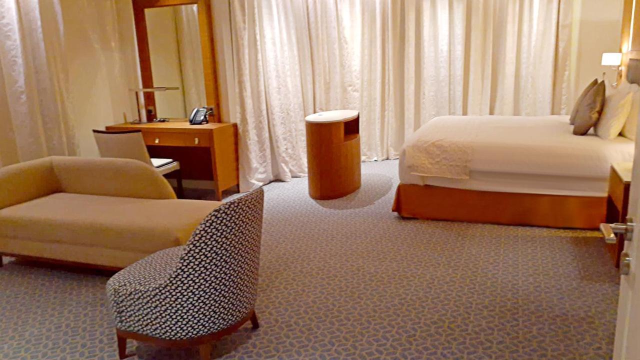 Grand Swiss-Belhotel Waterfront Seef Sanabis Esterno foto The photo shows a modern hotel room with a minimalistic design. There is a large bed against one wall with a simple headboard. Next to the bed, there is a small round table. On the opposite side, there is a comfortable-looking chair with a patterned 