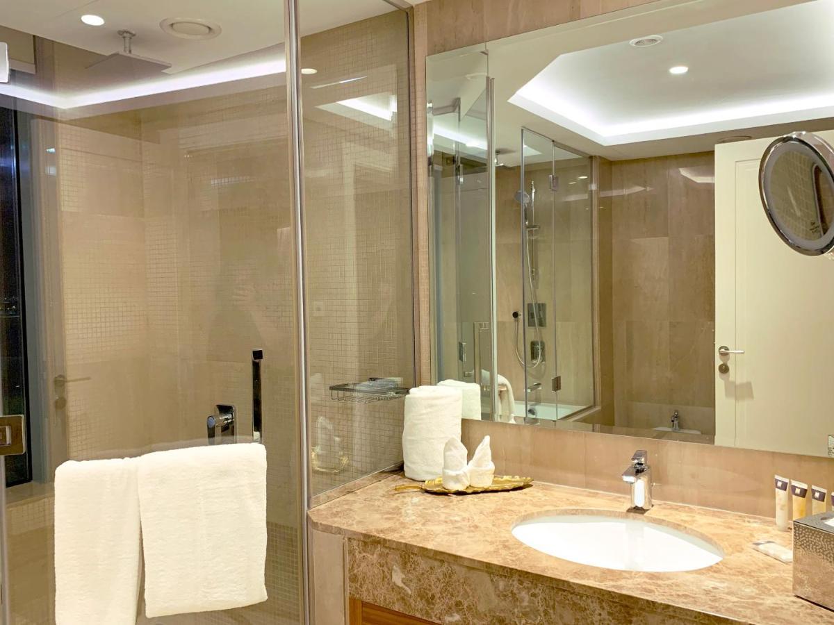 Grand Swiss-Belhotel Waterfront Seef Sanabis Esterno foto The photo shows a modern bathroom with a sleek design. There is a large mirror extending across the wall, reflecting the light fixtures above. The shower area is enclosed in glass, indicating a spacious shower. A marble countertop features a circular
