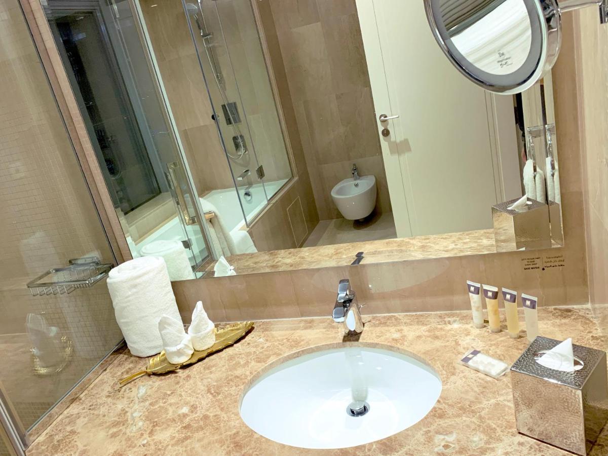 Grand Swiss-Belhotel Waterfront Seef Sanabis Esterno foto The photo shows a modern bathroom featuring a marble countertop with a circular sink and a faucet. There are neatly arranged toiletries on the countertop, including small bottles and disposable cups. A roll of toilet paper is visible, as well as a sm