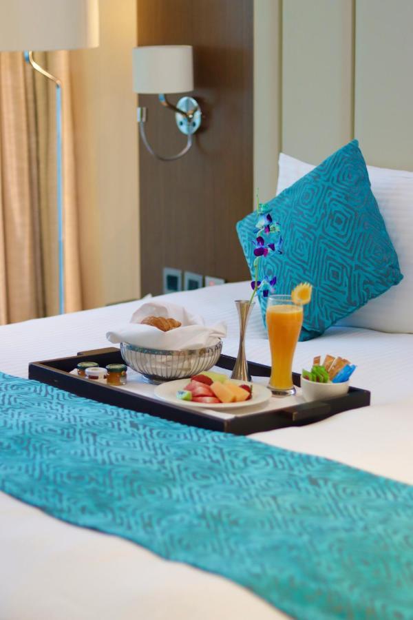 Grand Swiss-Belhotel Waterfront Seef Sanabis Esterno foto The photo shows a tray placed on a bed. On the tray, there is a variety of food items, including a basket with a pastry, sliced fruits, and small jars, likely containing condiments or spreads. There is also a tall glass filled with a bright orange be