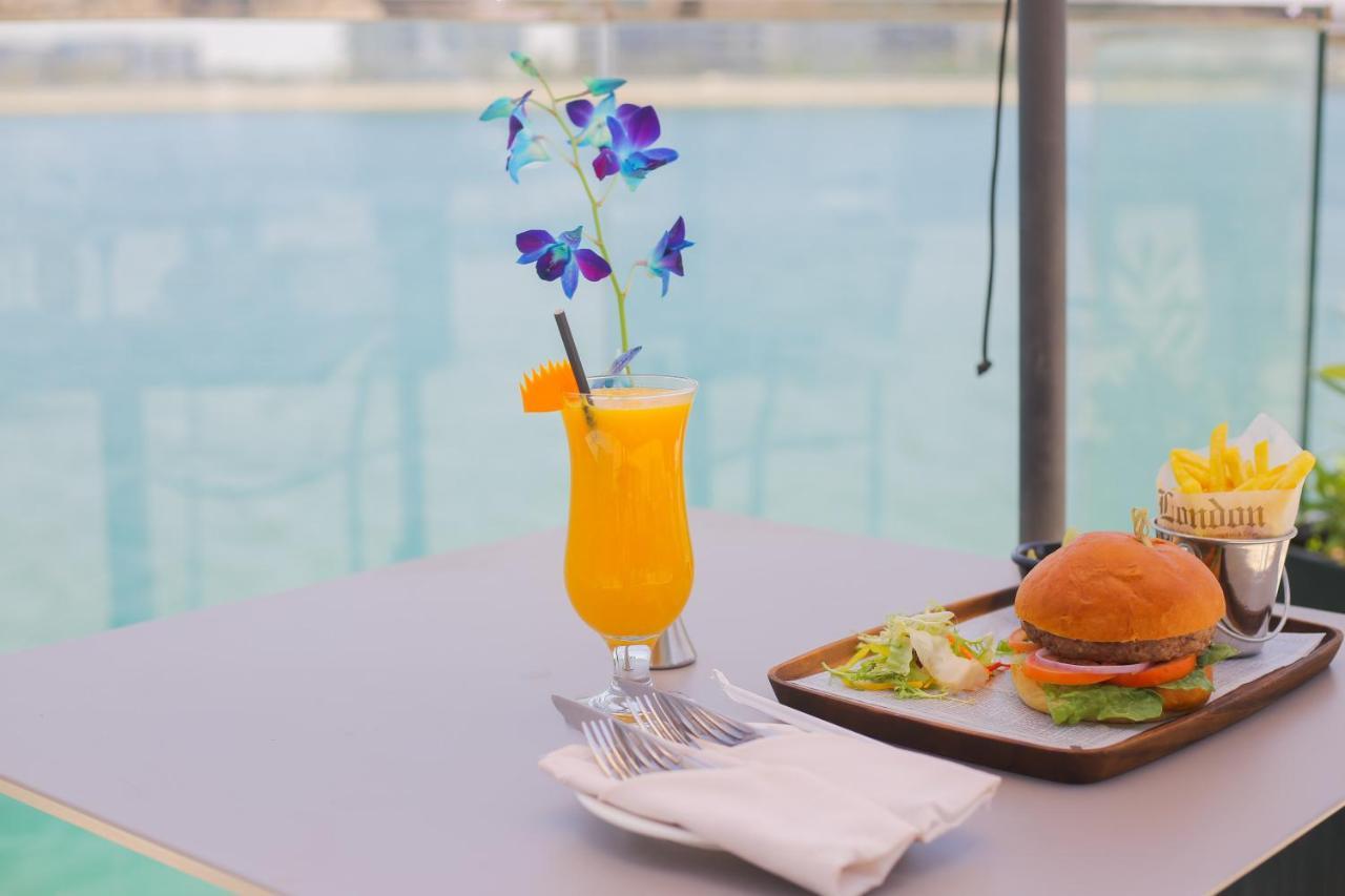 Grand Swiss-Belhotel Waterfront Seef Sanabis Esterno foto The photo features a refreshing scene with a vibrant orange drink garnished with a slice of orange and purple flowers, served in a glass. Next to it, there is a tray containing a hamburger with lettuce and possibly other condiments, accompanied by a 