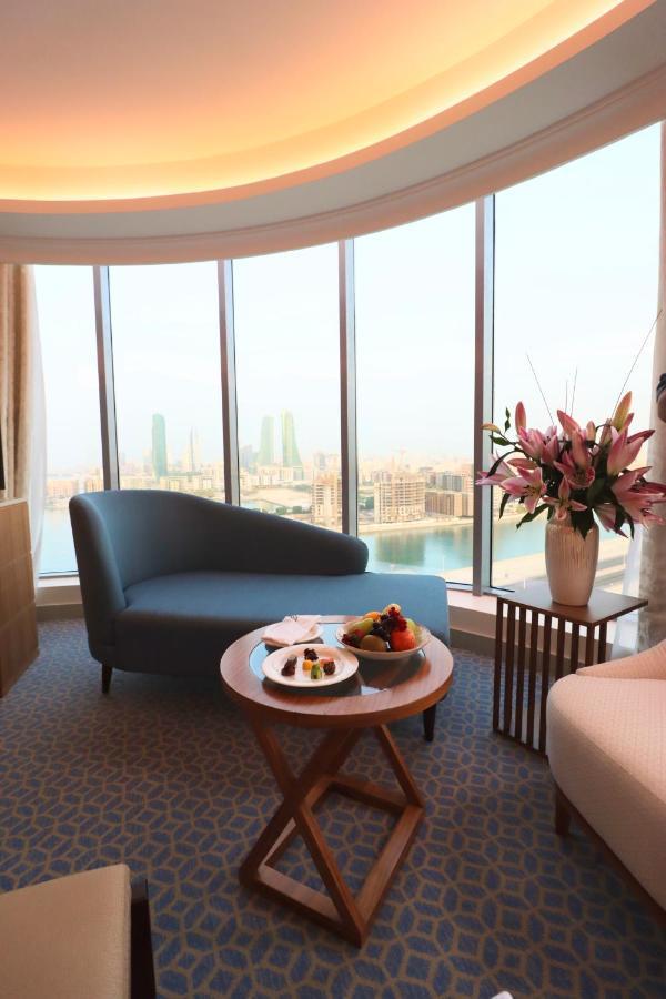 Grand Swiss-Belhotel Waterfront Seef Sanabis Esterno foto The image shows a stylish and modern interior of a hotel room or lounge area. There is a curved blue sofa positioned near large windows that offer a view of a cityscape, featuring tall buildings and what appears to be water in the foreground. A round