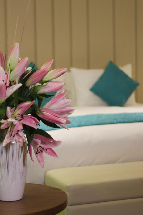 Grand Swiss-Belhotel Waterfront Seef Sanabis Esterno foto The image features a close-up of a bouquet of pink lilies positioned in the foreground, creating a vibrant and fresh atmosphere. In the background, there is a neatly made bed with white linens and a decorative turquoise pillow, adding a touch of colo