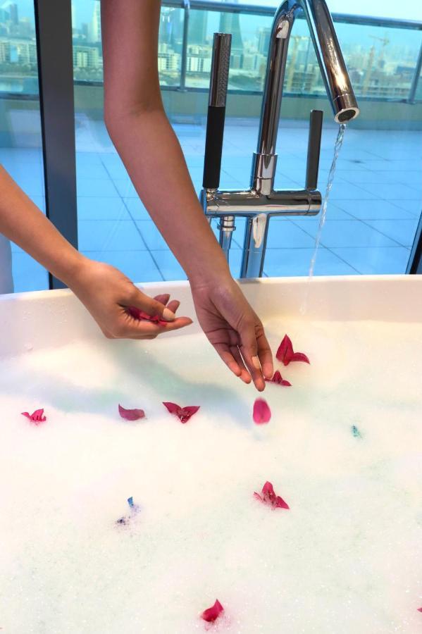 Grand Swiss-Belhotel Waterfront Seef Sanabis Esterno foto The photo shows a person's hand gently placing flower petals into a bath filled with bubbles. The bath appears to be modern and white, and the background suggests a clean, spacious environment. There is a faucet visible, and the overall scene evokes 