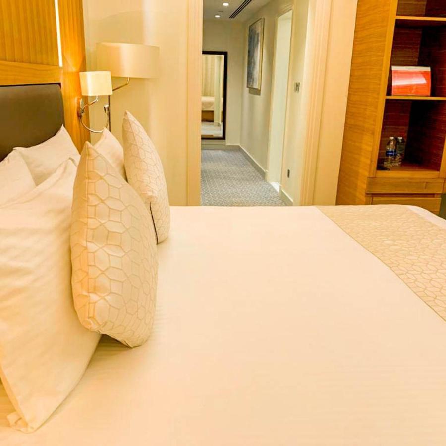 Grand Swiss-Belhotel Waterfront Seef Sanabis Esterno foto The photo shows a neatly made bed with a light-colored blanket and decorative pillows. In the background, there is a hallway leading to other room areas. The walls are painted in neutral tones, and there are elements of modern decor, such as a bedsid
