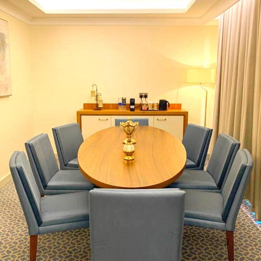 Grand Swiss-Belhotel Waterfront Seef Sanabis Esterno foto The photo shows a modern dining or conference room setup. It features a round wooden table surrounded by several blue upholstered chairs. In the center of the table, there is a decorative golden piece, likely a vase or trophy. To one side of the room