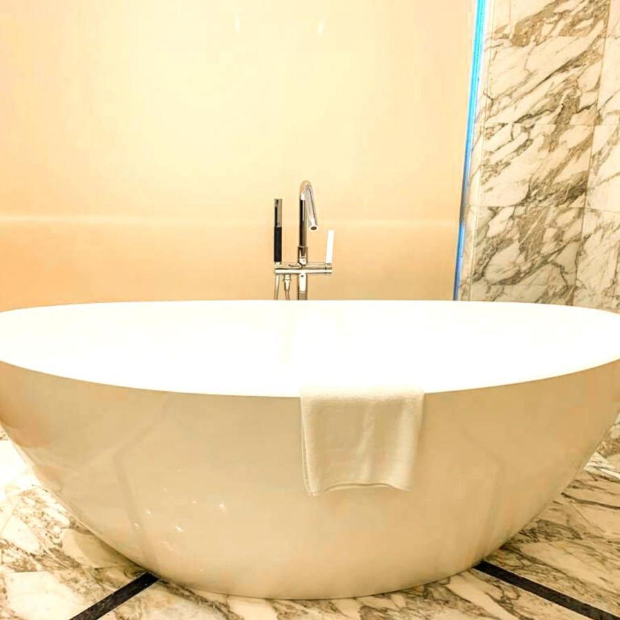 Grand Swiss-Belhotel Waterfront Seef Sanabis Esterno foto The photo shows a modern, oval-shaped bathtub located in a bathroom. The tub is white and has a sleek, minimalist design. A chrome faucet with a handle is mounted above the tub. There’s a white towel draped over the edge of the tub. The surrounding w