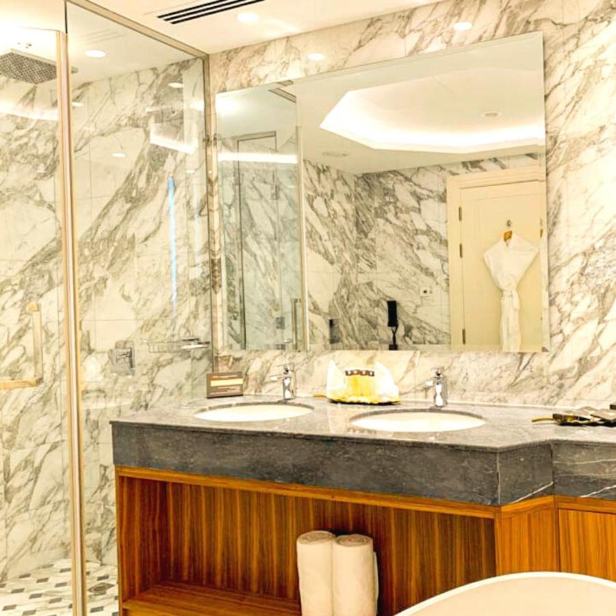 Grand Swiss-Belhotel Waterfront Seef Sanabis Esterno foto The photo depicts a luxurious bathroom featuring elegant marble walls. There are two sinks with a dark granite countertop, and the cabinetry below has a warm wooden finish. Large mirrors are mounted above the sinks, reflecting the stylish design. A w